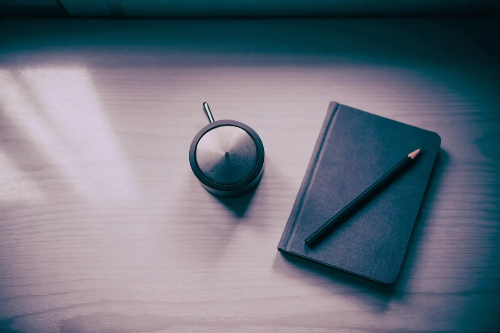 A small round kitchen timer and a closed notebook with a pencil on a wooden desk, photographed from above in soft window light. Nobody is at the desk.