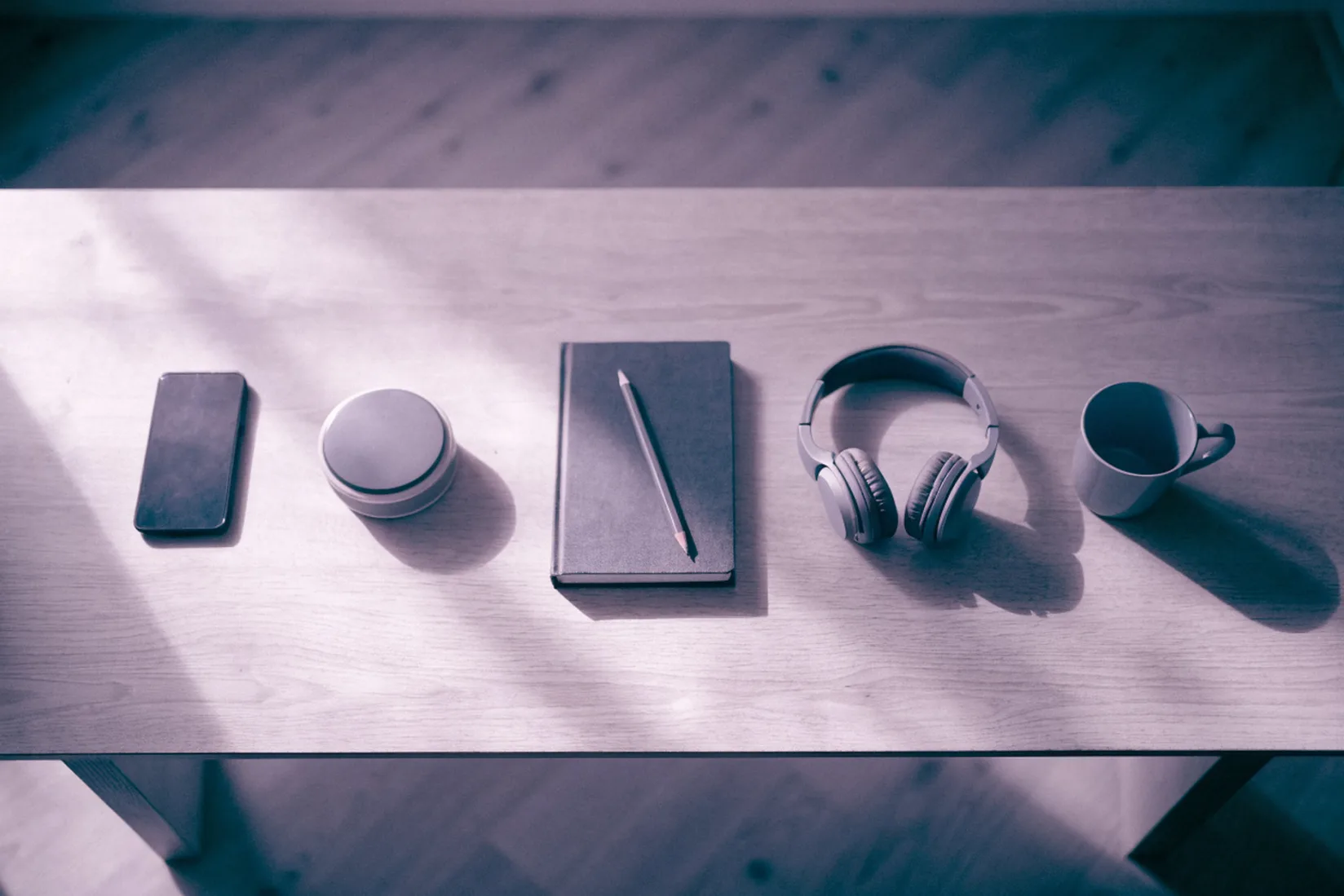 Five everyday objects in a neat row on a wooden desk seen from above: a phone, a small round timer, a notebook with a pencil, a pair of headphones and a mug, in soft window light.