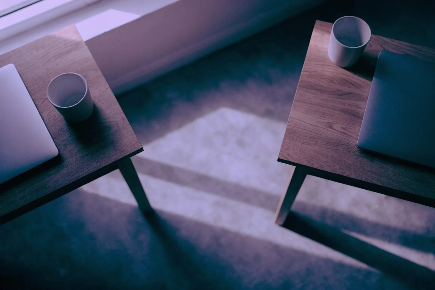 Two small wooden desks seen from above, each holding a closed laptop and a plain white cup, with a strip of soft window light falling on the floor between them.